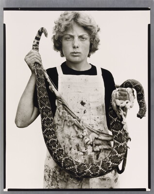 A black-and-white portrait photograph of a White teenage boy with curly hair wearing a dirty apron and holding a gutted snake in front of him.