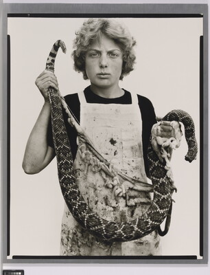 A black-and-white portrait photograph of a White teenage boy with curly hair wearing a dirty apron and holding a gutted snake in front of him.