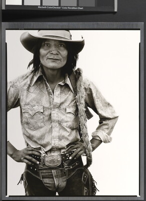 A black-and-white portrait photograph of a Native American man with long hair wearing a cowboy hat, button-down shirt, jeans, and chaps, standing with hands on hips.
