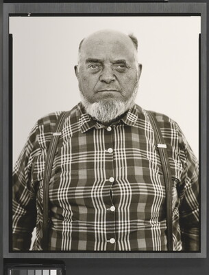A black-and-white portrait photograph of an older, balding White man with a gray beard wearing a plaid button-down shirt and suspenders.