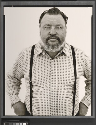 A black-and-white portrait photograph of a middle-aged White man with a receding hairline and salt-and-pepper beard wearing a checked button-down shirt and suspenders.