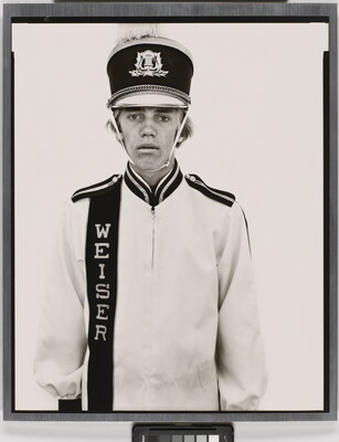 A black-and-white portrait photograph of a White teenage boy wearing an oversized, white, marching-band hat and uniform.