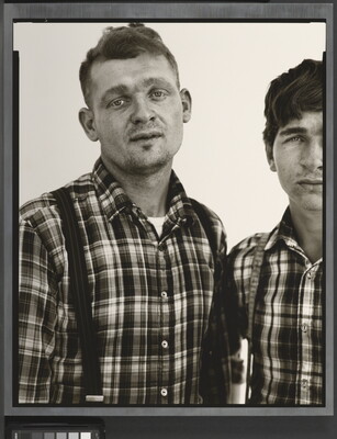 A black-and-white portrait photograph of two White men, both in similar plaid shirts and suspenders, standing shoulder to shoulder.