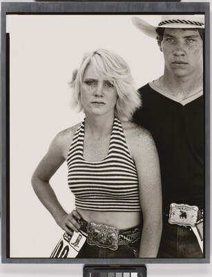 A black-and-white portrait photograph of a young White woman wearing a striped halter top, one hand on her hip, and a young White man wearing a cowboy hat and V-neck t-shirt, both wearing large buckles on their belts.