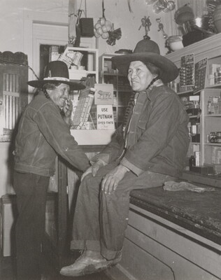 A black-and-white photograph of two Native American people in a general store: one wearing a jean jacket and cowboy hat sitting on a counter and the other dressed similarly standing nearby.