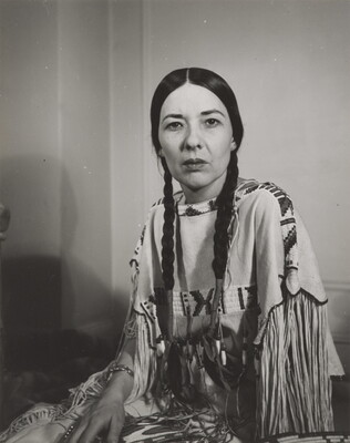 A black-and-white photograph of a seated Indigenous woman, hair in braids, gazing into the camera. 