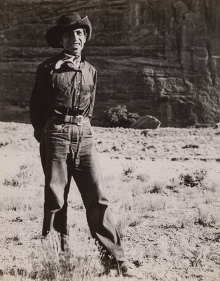 A black-and-white photograph of a man wearing a cowboy hat and neckerchief posing in front of what looks like the face of a cliff.