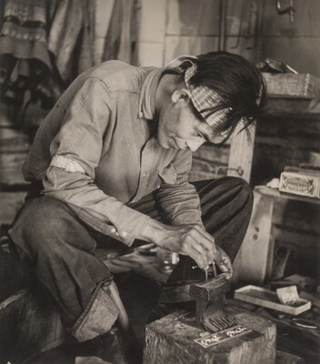 A black-and-white photograph of a seated Native American man bent over an anvil in a workshop.