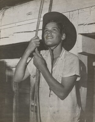A black-and-white photograph of a medium-skinned boy in a cowboy hat holding a couple of ropes suspended from above.