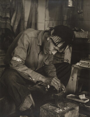 A black-and-white photograph of a seated Native American man bent over an anvil in a workshop.