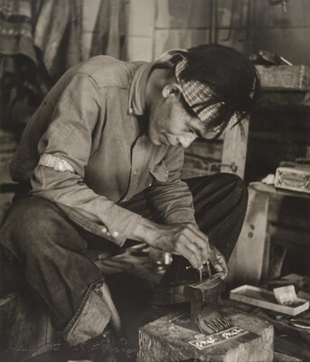 A black-and-white photograph of a seated Native American man bent over an anvil in a workshop.