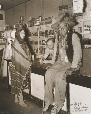 A sepia-toned photograph of a Indigenous man sitting on a counter, an Indigenous woman wrapped in a blanket standing at the counter, and a White man behind the counter.