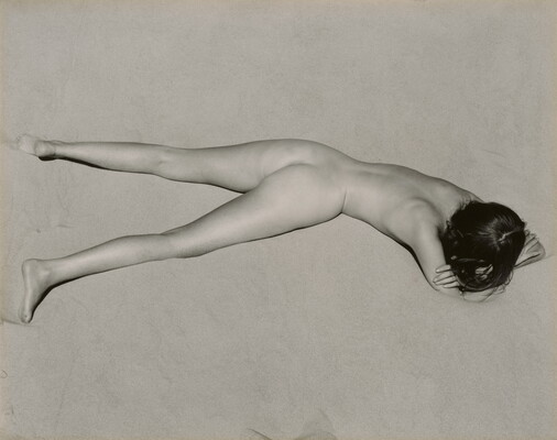 A black-and-white photograph of a nude, fair-skinned woman lying facedown in the sand with her head resting on her crossed arms.
