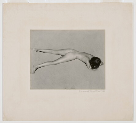 A black-and-white photograph of a nude, fair-skinned woman lying facedown in the sand with her head resting on her crossed arms.