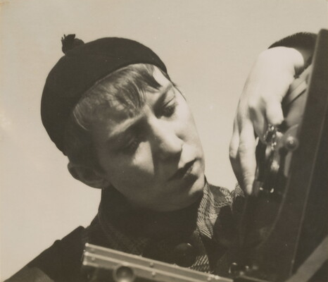 A black-and-white photograph of a White woman with short hair, wearing a beanie hat, manipulating a large format camera.