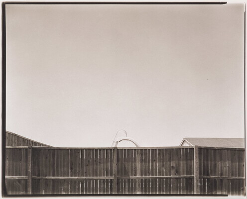 A black-and-white photograph of a wood privacy fence; the top of a pool slide and a roof are seen just above it.