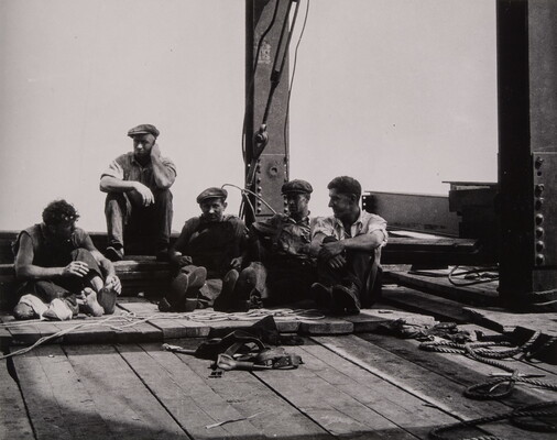 A black-and-white photograph of five men sitting on wood planks in between steel beams on a construction site.