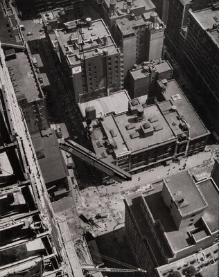 A black-and-white photograph looking down on the buildings of a city and several steel girders suspended by cables in mid-air.