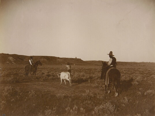 A black-and-white photograph of two men on horses watching as a third man ropes a cow.