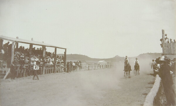 A black-and-white photograph of people on running horses coming around a bend in a dirt racetrack with spectators in stands on either side.