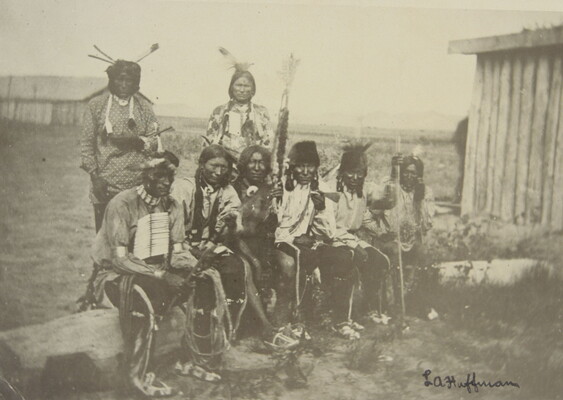 A black-and-white photograph of six Native Americans sitting on a log and two standing behind them, all wearing traditional clothing.