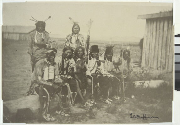 A black-and-white photograph of six Native Americans sitting on a log and two standing behind them, all wearing traditional clothing.