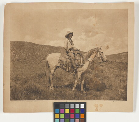 A sepia-toned photograph of a cowboy on a horse standing in a hilly plain.