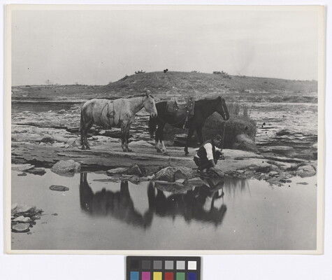 A black-and-white photograph of a man crouched down dipping his hat into a body of water while two horses stand behind him.