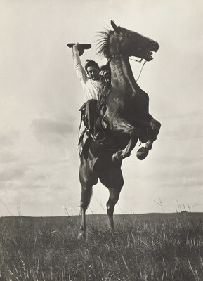 A black-and-white photograph of a White man on a rearing horse holding his hat in one hand above his head.