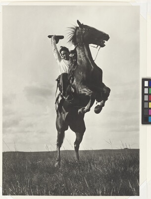 A black-and-white photograph of a White man on a rearing horse holding his hat in one hand above his head.