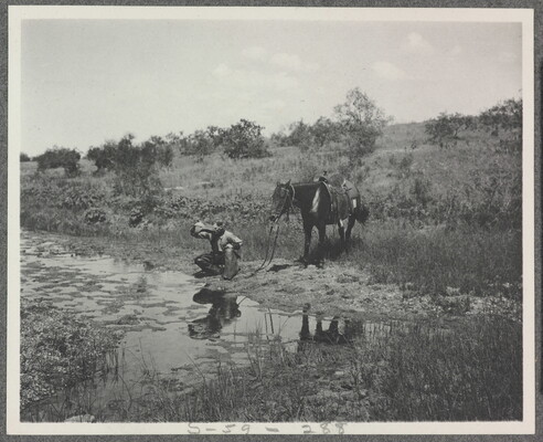 A black-and-white photograph of a cowboy crouched next to a stream drinking water from his hat as his horse stands behind him.