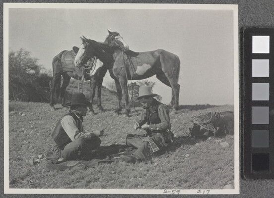A black-and-white photograph of two cowboys playing a card game on the ground, while their horses stand nearby.