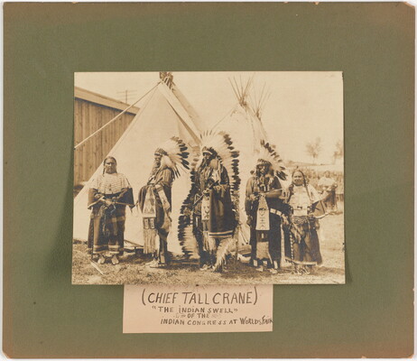 A sepia-toned photograph mounted on green board of a group of Native Americans in full regalia in front of tipis.