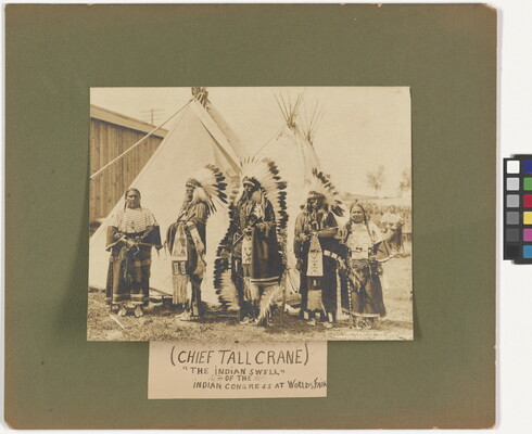 A sepia-toned photograph mounted on green board of a group of Native Americans in full regalia in front of tipis.