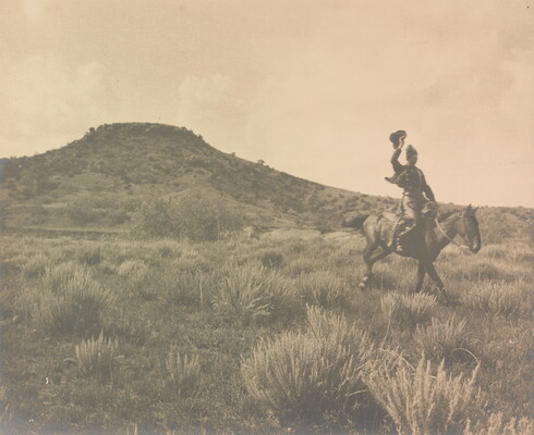 A black-and-white photograph of a person on horseback, one arm thrown up with hat in hand, in a scrubby terrain.