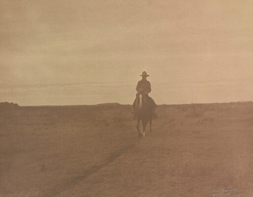 A sepia-toned photograph of a cowboy on horseback trotting on a path toward the viewer in a barren landscape.