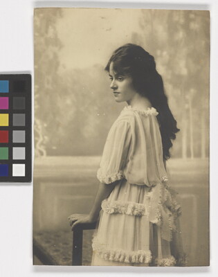 A black-and-white photograph of a young White woman with long dark hair wearing a ruffled dress standing back to the camera but head turned in profile.