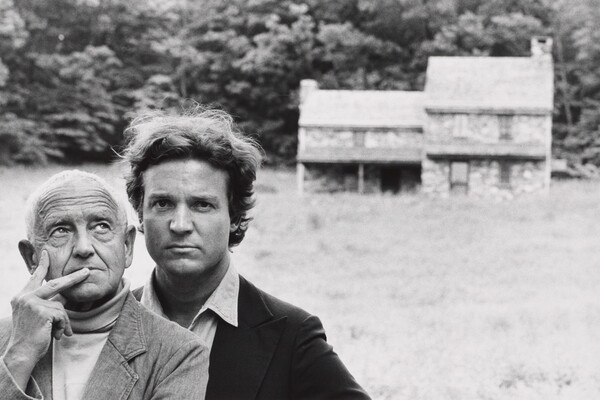 A black-and-white photograph of two White men, one older and one younger, standing in front of a stone farmhouse in the distance.