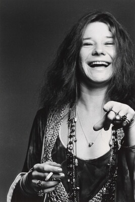 A black-and-white portrait photograph of a White woman with long hair, an open-mouthed smile, holding a cigarette in one hand and pointing at the camera with the other.