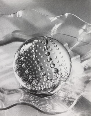 A black-and-white still life photograph of a clear glass, circular paperweight filled with bubbles in a clear, wavy glass bowl on a table.