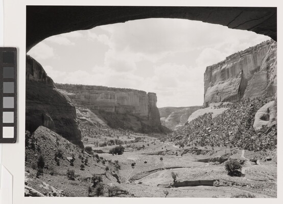 A black-and-white photograph of a canyon landscape framed by an arched rock overhang.