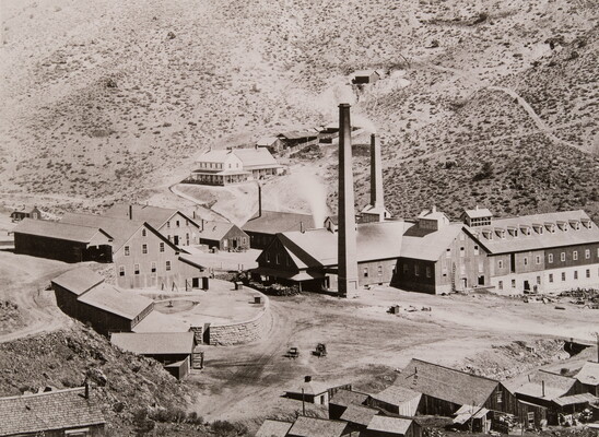 A black-and-white photograph of industrial buildings with smoke stacks in a rocky valley.
