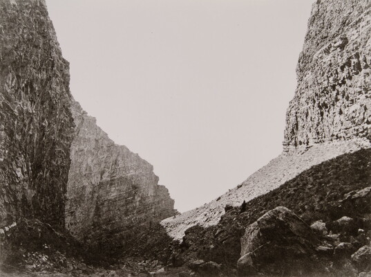 A black-and-white photograph of steep and rocky cliffs rising on both sides of a rocky valley.