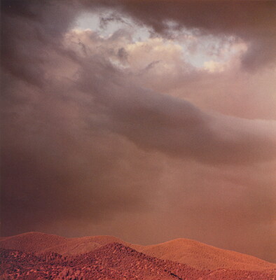 A color photograph of reddish mountain tops under dark, heavy clouds.