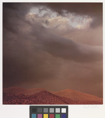 A color photograph of reddish mountain tops under dark, heavy clouds.