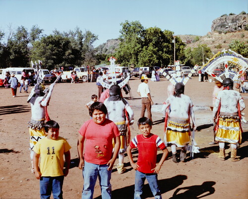 A color photograph of a three boys posing as a Native American dance takes place behind them.