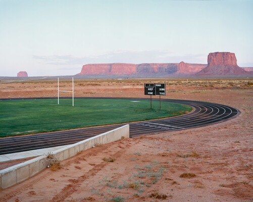 A color photograph of an athletic track and field with sunlit mesas and buttes in the distance.