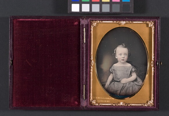 A color photograph in a gold, oval-shaped frame of a toddler-age White child with curled hair in a black-and-white checked dress.