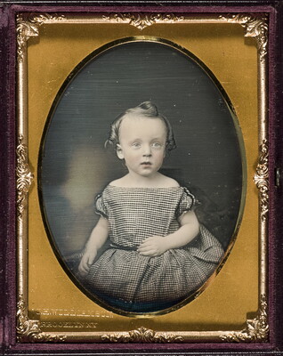 A color photograph in a gold, oval-shaped frame of a toddler-age White child with curled hair in a black-and-white checked dress.