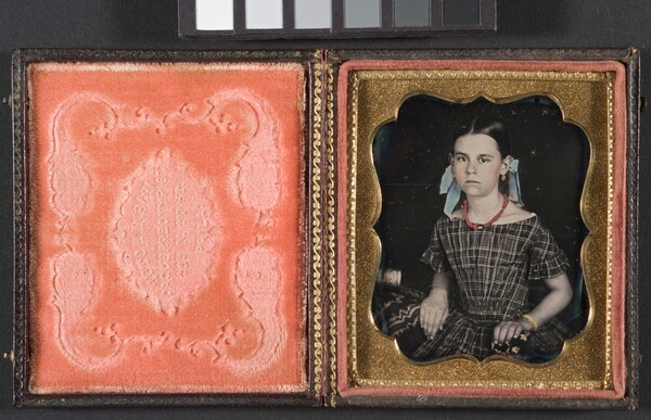 A hand-colored photograph in a decorative gold frame of a young White girl seated, wearing a plaid dress and bows in her hair.